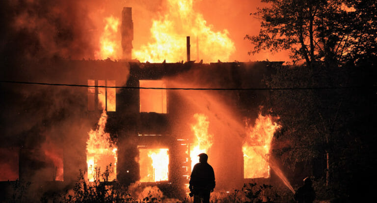 A fire being fought by fire fighters with a man's silhouette in-front of the burning building.