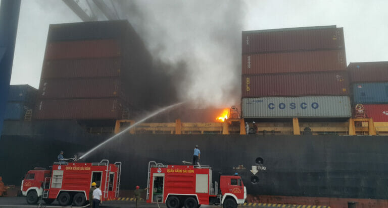 A cargo ship and shipping containers on fire with fire fighters on scene